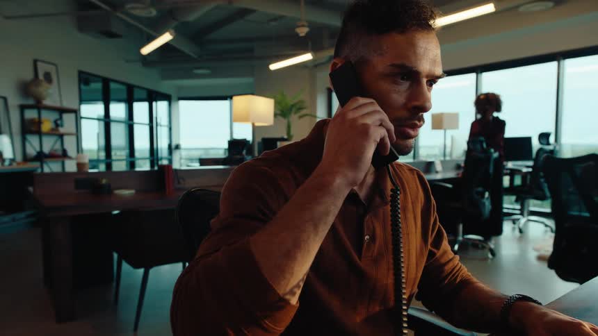 A focused businessman types on his computer before answering a landline telephone call in a modern, open-concept office