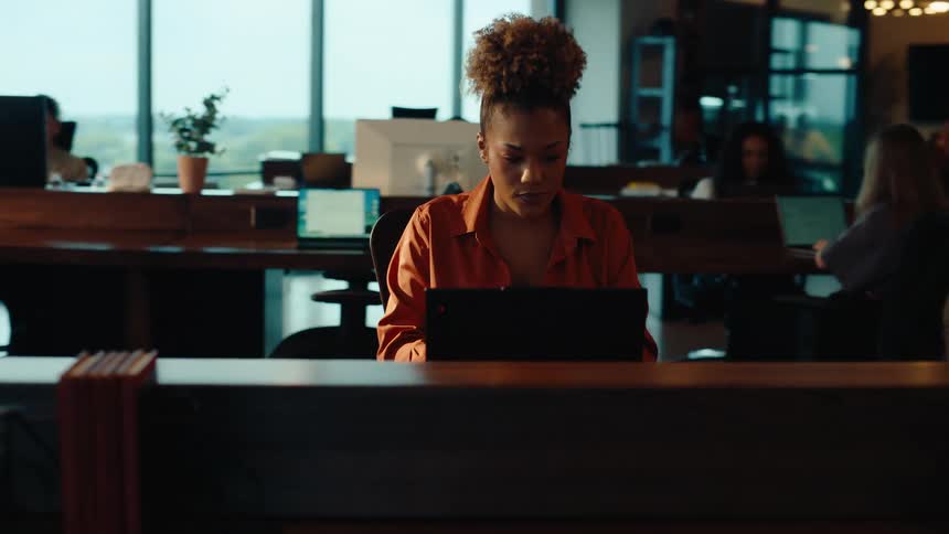 A focused young African American woman diligently works on her laptop in a bustling, modern open-plan office