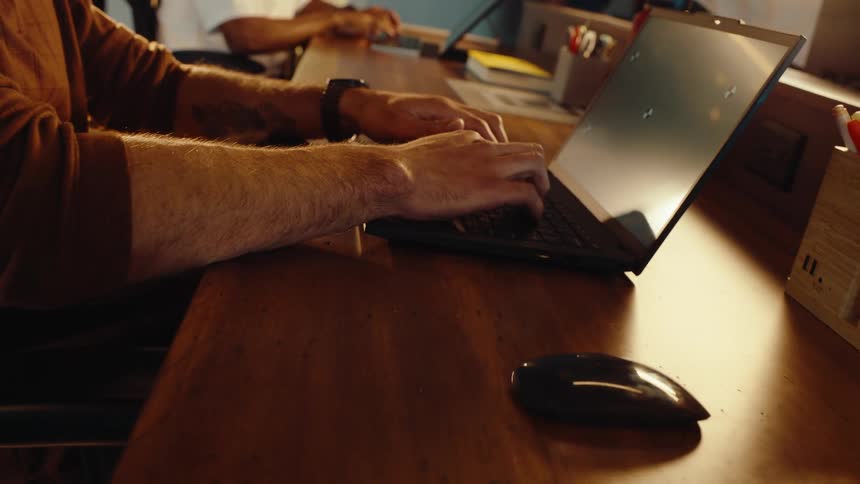 A close-up shot pulls back from a man's hands with a tattoo typing on a laptop with tracking markers on the screen in a modern office