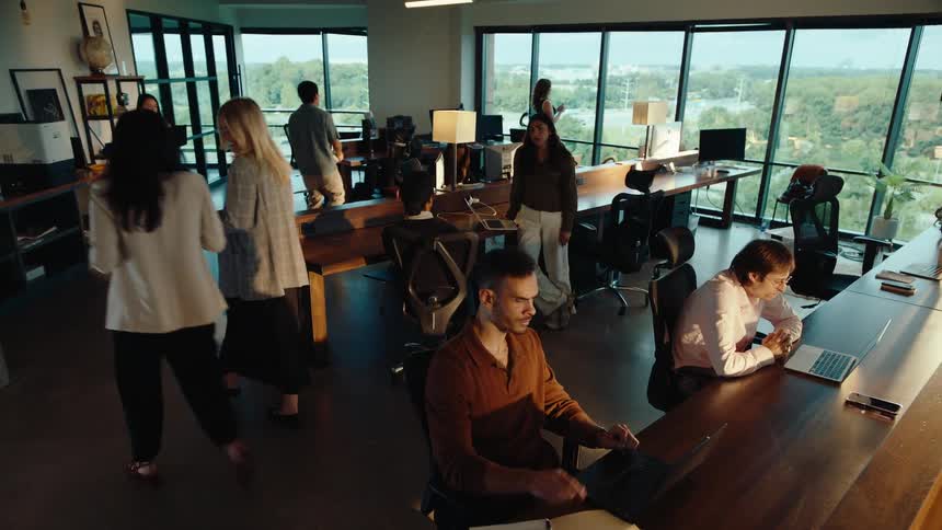 Crane shot rises from a focused young man working on a laptop to a wide view of a busy, modern open-plan office at sunset