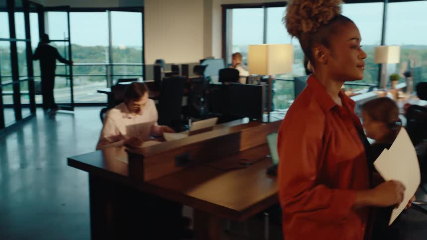 A professional woman walks through a bustling, modern open-plan office to deliver a file to a colleague at his desk