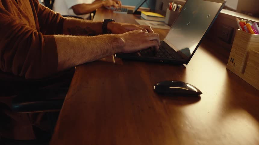 A slow push-in shot focuses on a man's hands as he types on a laptop with a green screen while sitting at his desk