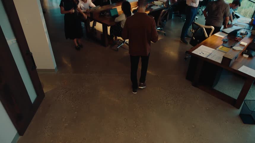 From a high-angle view, a businessman walks through a bustling, modern open-plan office to join his colleagues