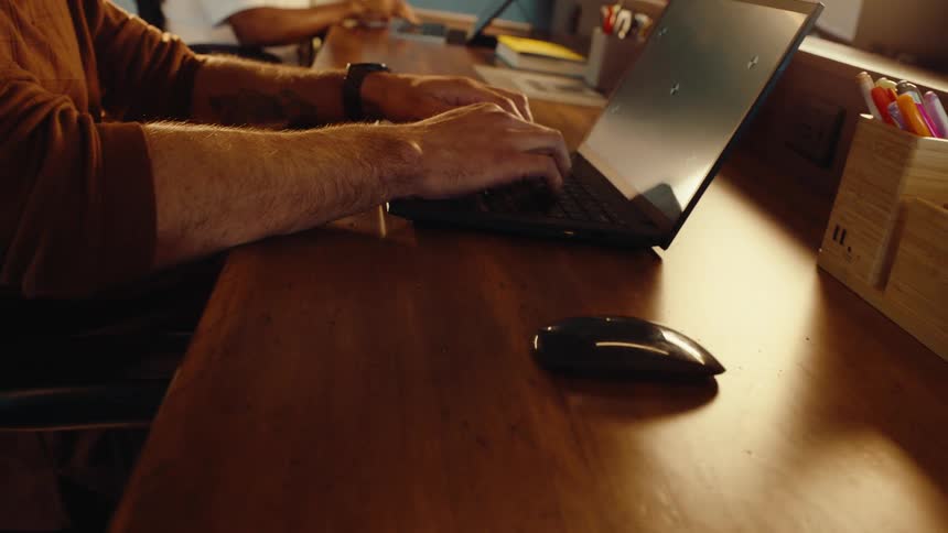 A dolly-out shot of a man's hands typing on a laptop with a greenscreen at a warmly lit wooden office desk