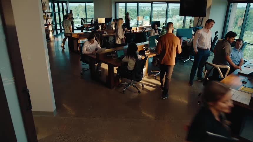 Overhead crane shot follows a man walking into a bustling, modern open-plan office, revealing a collaborative team at work
