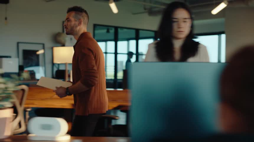 A male employee walks through a modern open-plan office in slow motion to collaborate with a female colleague at her desk