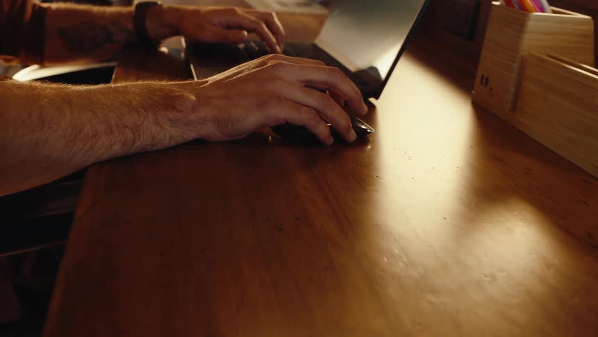Close-up dolly shot of a man's tattooed arm and hand using a wireless mouse while working on a laptop at a modern wooden desk