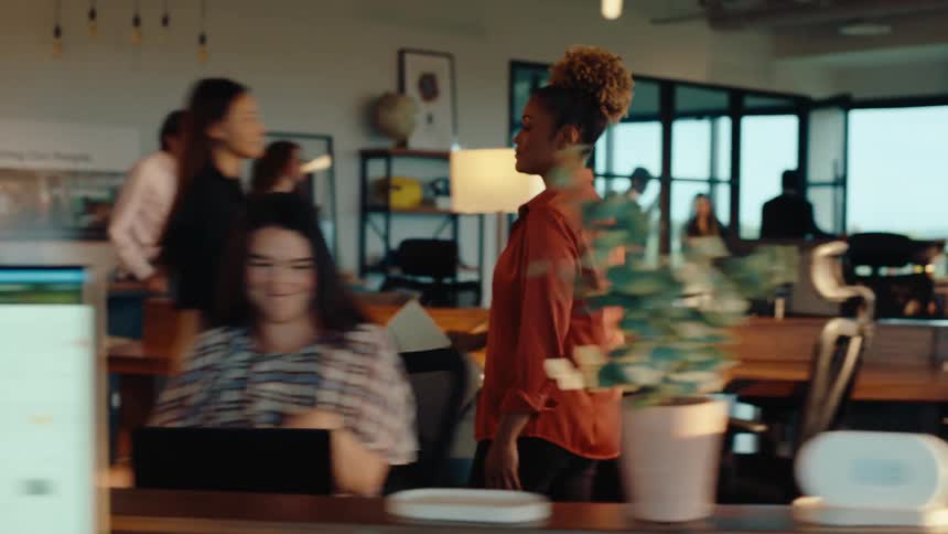 A professional woman walks through a busy, modern office in slow motion to collaborate with a coworker at her desk