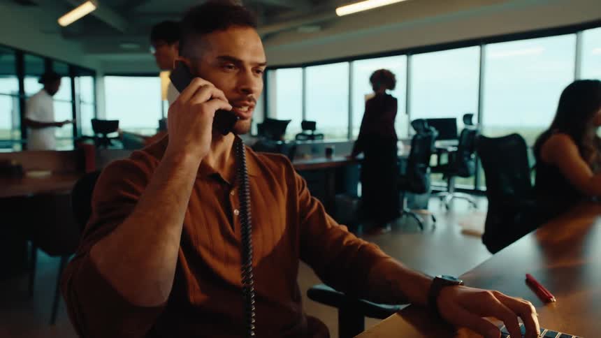 A young businessman types on his keyboard and answers a landline phone call while working at his desk in a modern office