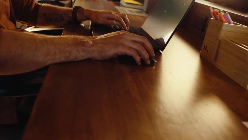Slow pullback shot of a man's hands typing on a laptop and using a mouse at a wooden desk in a warmly lit office
