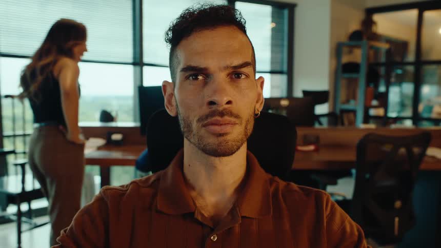 A young male employee types at his desk before quickly looking up to face the camera with a serious expression in a modern office