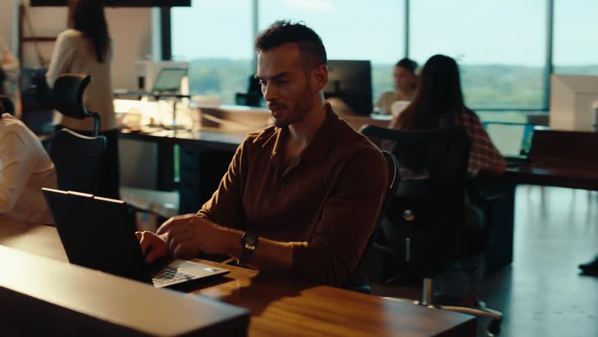 A focused businessman works diligently on his laptop in a modern, open-plan office environment