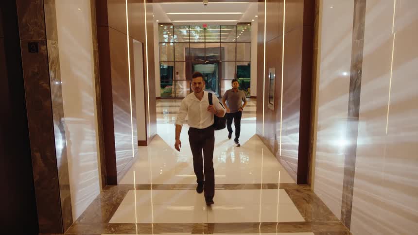 A steady flow of professionals make their way through a modern corporate hallway towards the elevators in slow motion