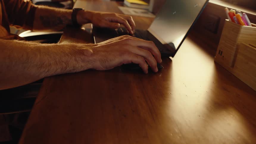 Slow push-in shot captures a man's hands typing and using a mouse on a laptop at a warm wooden desk