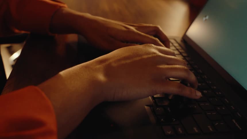 Close-up dolly shot of a person's hands typing on a laptop keyboard with a green screen in a warmly lit room