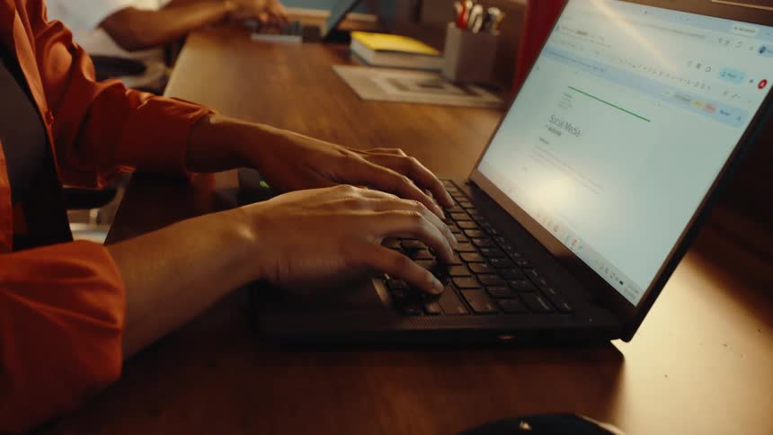 A close-up shot of a professional typing on a laptop at a wooden desk in a modern, warmly lit office