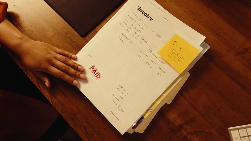 Top-down view of a person's hands stamping an invoice with a red 'PAID' stamp at a wooden desk