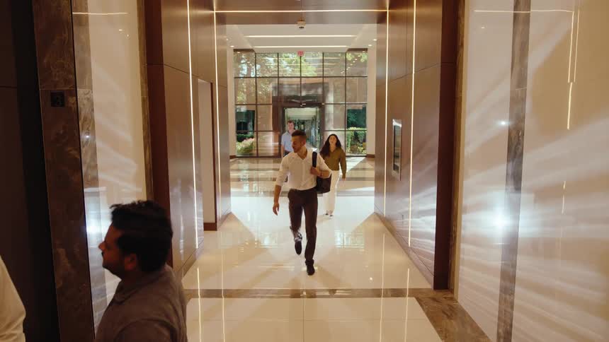 A diverse group of business professionals walks through a modern office building lobby towards an elevator in slow motion