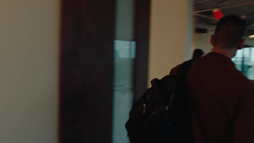 A young professional carrying a laptop walks through a stylish hallway and enters a bustling, open-plan office to join his colleagues at work