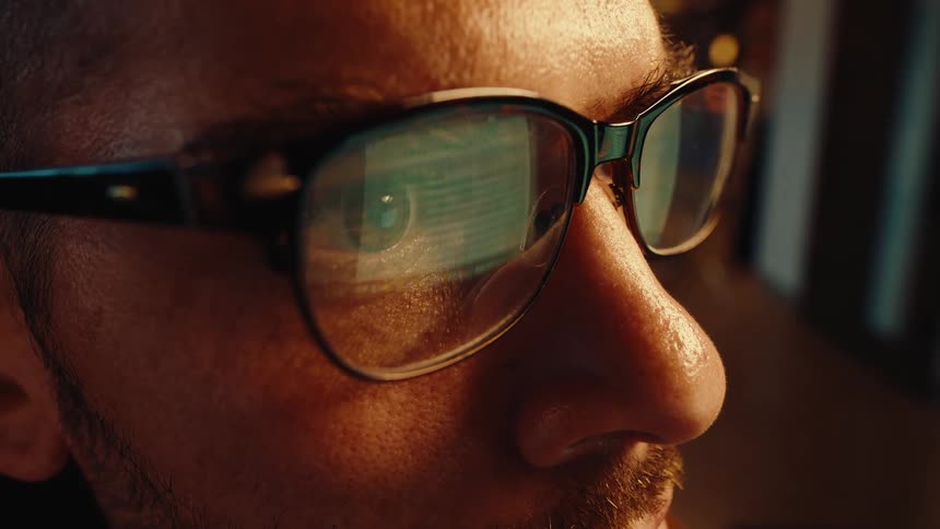 An extreme close-up cinemagraph of a man's face in profile with a computer screen reflecting in his glasses as he works