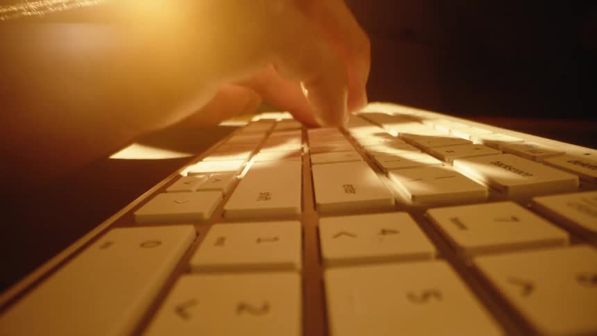 A cinematic slow dolly shot captures a hand typing on a white computer keyboard illuminated by a warm golden light in a dark room