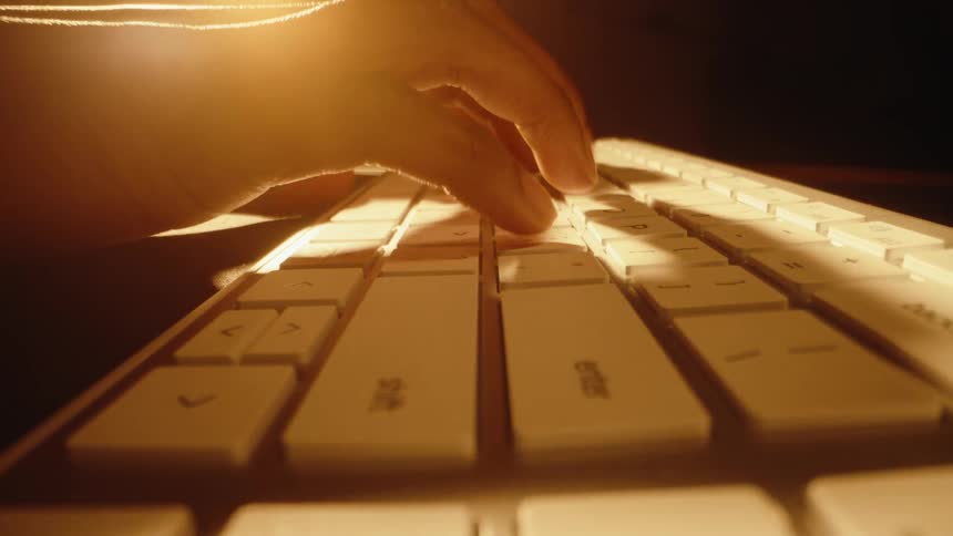 An extreme close-up of a hand typing on a modern white keyboard, illuminated by a warm, golden light in a dark setting