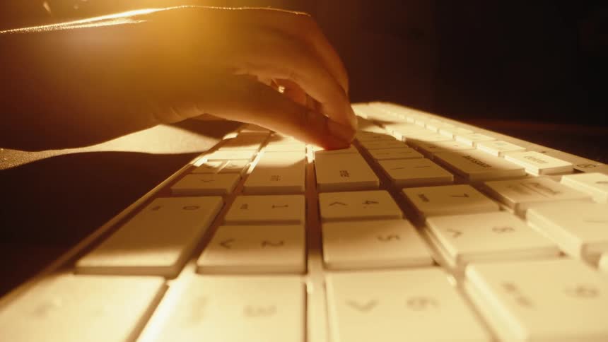 A dramatic, slow push-in close-up shot of a person's hand typing on a white keyboard in a dark, warmly lit room