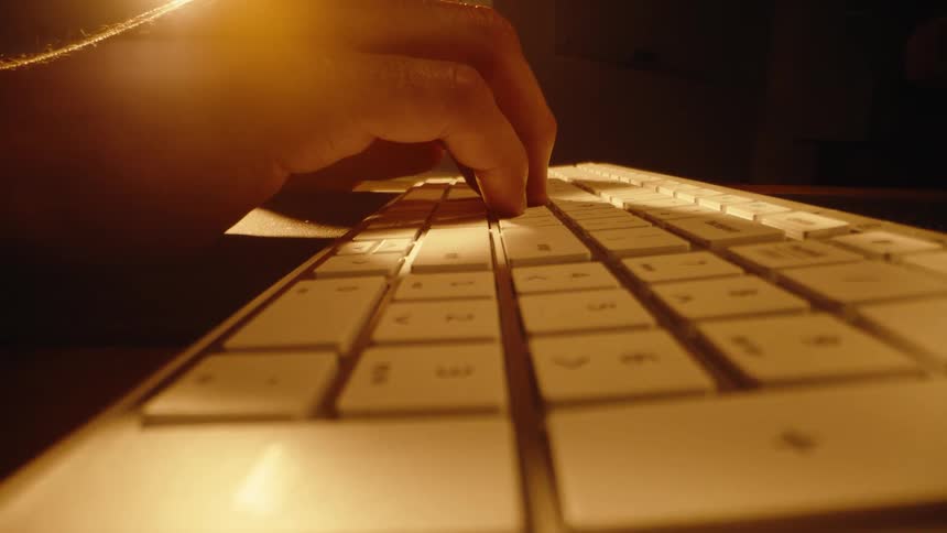 A stylistic, low-angle close-up of a hand typing on a computer keyboard in a dark room with warm lighting