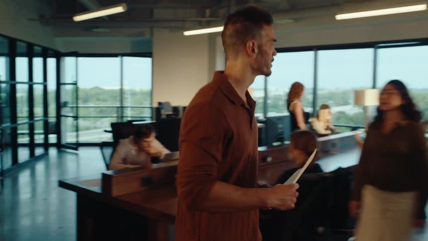 A male employee confidently walks through a busy modern office in slow motion to hand important documents to a colleague at their desk
