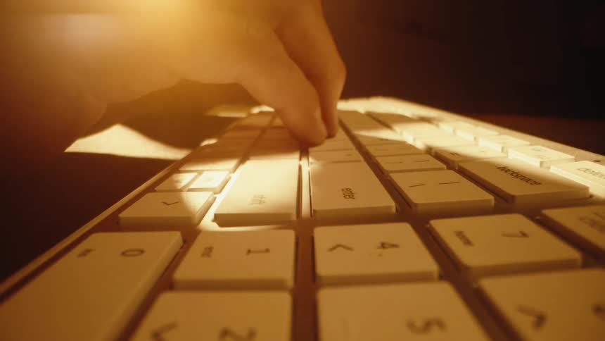 A dramatic close-up dolly shot of fingers typing on a white computer keyboard in warm, moody lighting