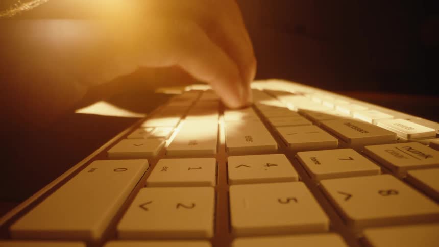 A dramatic slow pull-back shot of a person's fingers typing on a modern computer keyboard in a warmly lit, dark room
