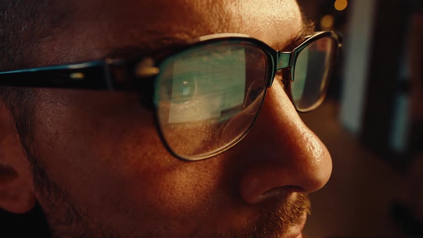 Close-up of a programmer's glasses reflecting scrolling code on the screen as he works late at night