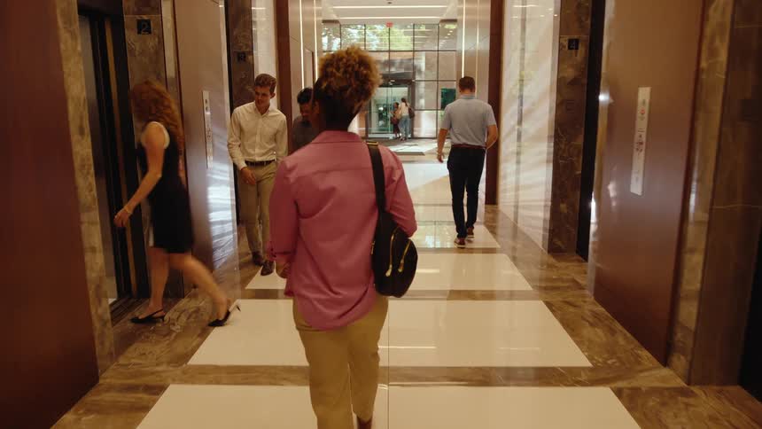 Following a young Black woman from behind as she walks through a modern office hallway and steps into an elevator with her coworkers