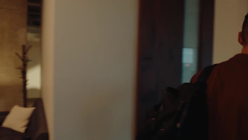 A young professional carries his laptop and backpack as he walks through a modern open-plan office filled with coworkers