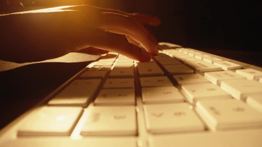 A dramatic, slow-motion pull-back shot reveals fingers typing on a white keyboard in a dark room illuminated by warm light