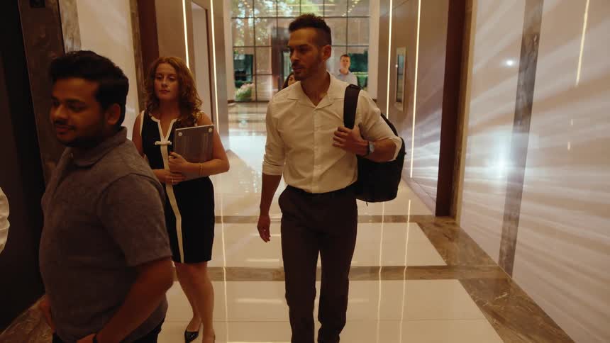 Diverse group of business professionals walk down a modern hallway and enter an elevator together, laughing and talking before the doors close