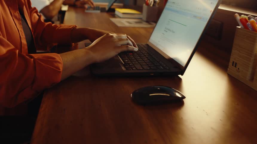 A slow push-in shot captures the hands of a professional typing on a laptop at a wooden desk in an office