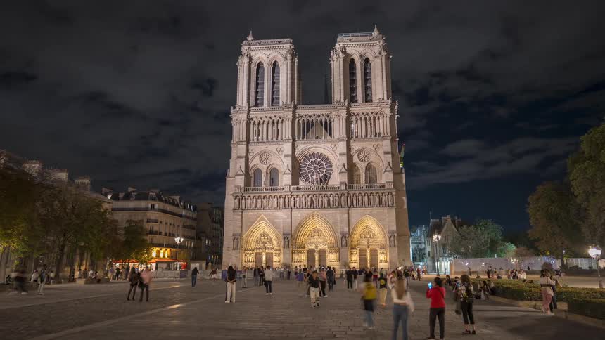 A dramatic nighttime timelapse pulling back from the illuminated facade of the historic Notre-Dame Cathedral in Paris as crowds of people wander through the plaza