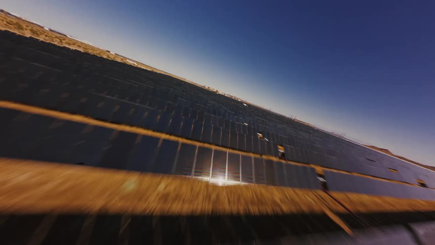 A dynamic FPV drone flies at high speed just above the rows of a massive solar panel farm in the desert