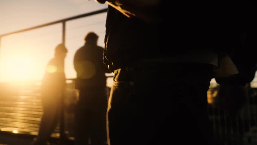 A slow-motion close-up of a person's silhouette by a fence during a golden sunset, with others in the background