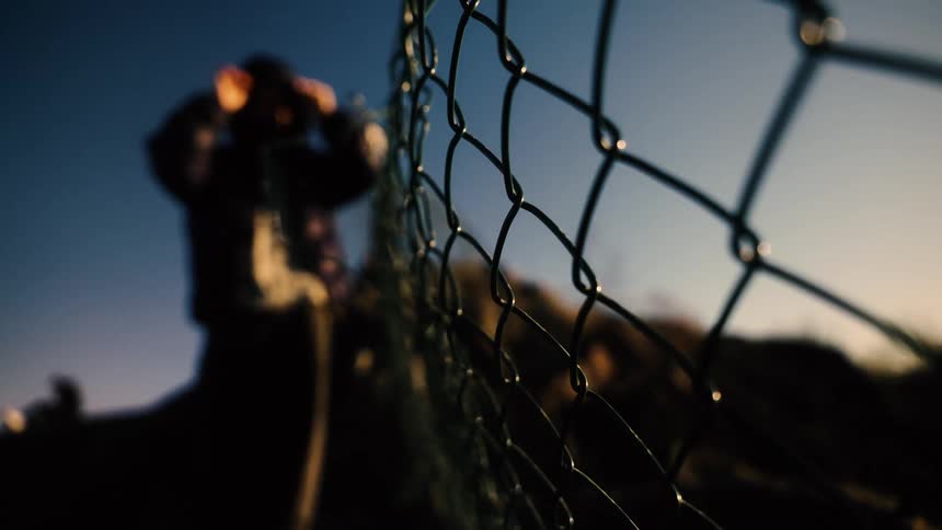 A mysterious hooded figure stands behind a chain-link fence at sunrise in this dramatic, low-angle shot