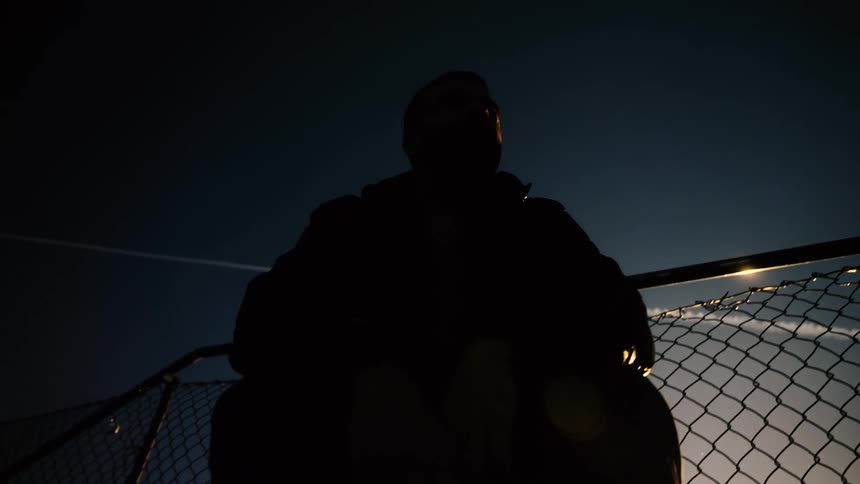 A solitary man is silhouetted against the dark blue twilight sky as he leans on a chain-link fence