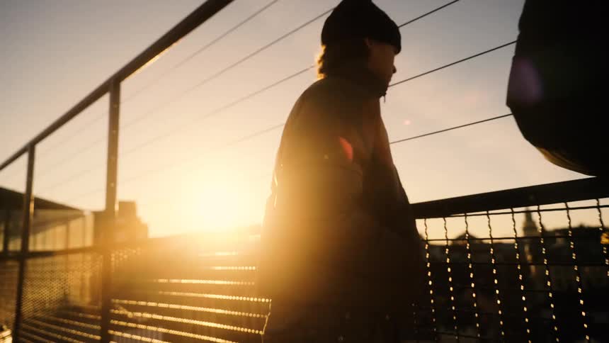 A person wearing a jacket walks along a chain-link fence and then stops on a bridge to watch the beautiful golden sunset over the city
