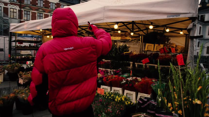 Following a person in a striking red puffer jacket as they browse a vibrant flower stall at a city market