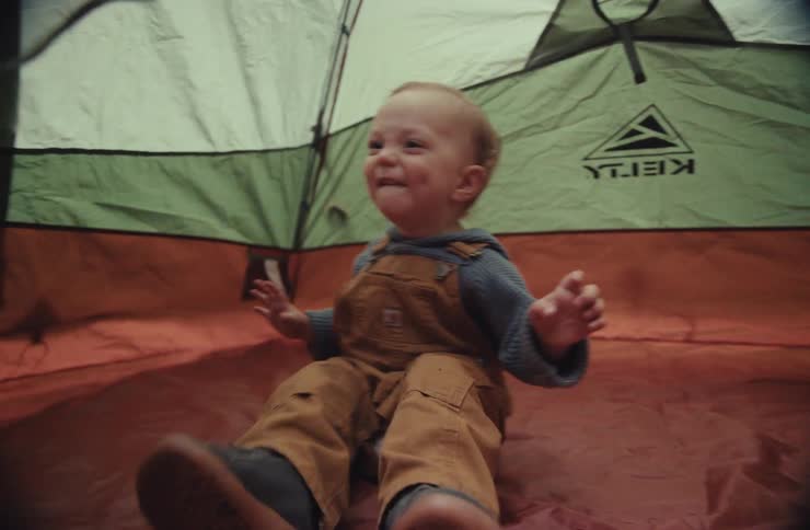 Adorable toddler in overalls happily waves and kicks his feet in slow motion while sitting inside a camping tent