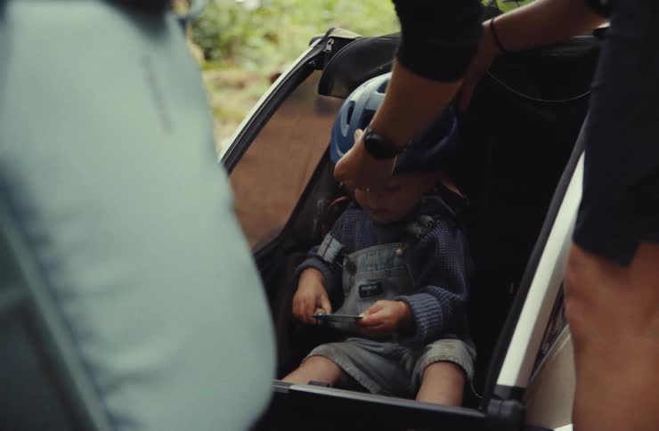 Parents carefully put a bicycle helmet on their happy toddler who is sitting in a child carrier trailer