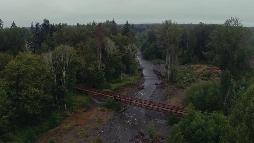 An aerial shot slowly moves in as two cyclists ride their mountain bikes across a bridge over a rocky river in a dense forest