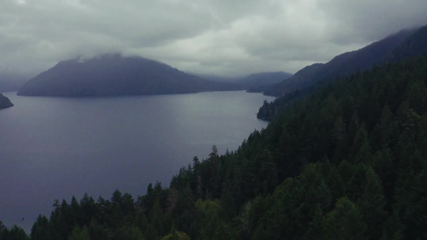 An aerial view flies over a dense evergreen forest revealing a vast, serene lake surrounded by mountains on a cloudy day