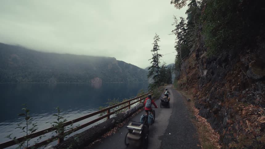 Two adventurers bike packing down a paved path next to a misty mountain lake on an overcast day