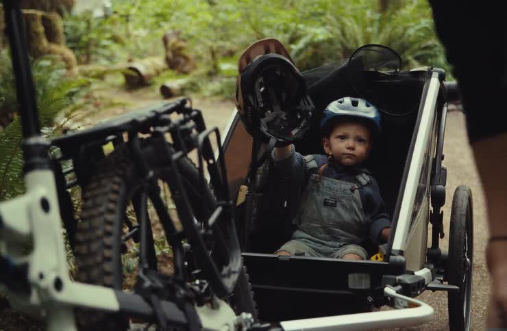 A parent packs a bag for an adventure while their young child sits in a bike trailer in the forest, holding up a helmet